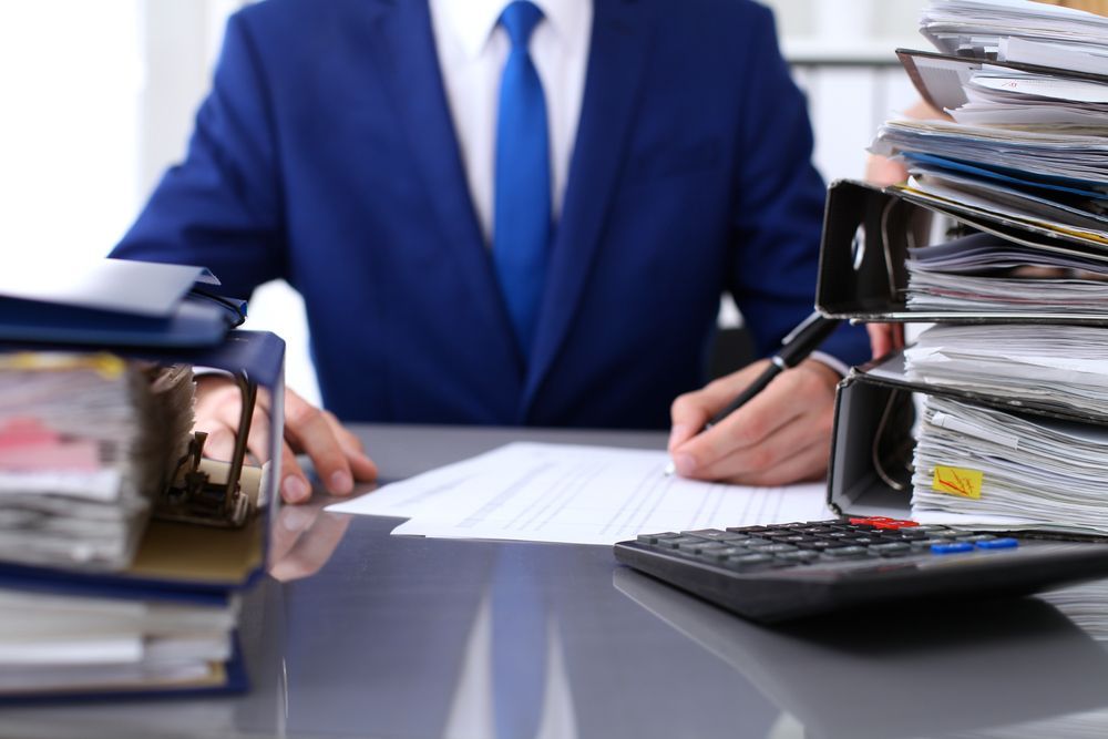 Man in suit reviewing documents at a desk with a calculator, surrounded by files.