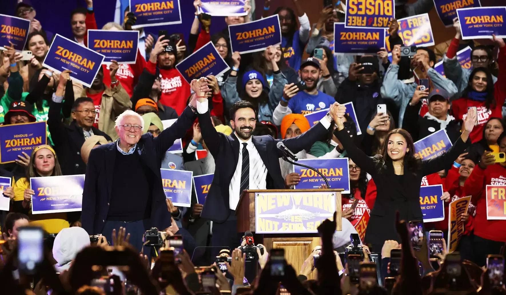 Zohran Mamdani, Bernie Sanders, and Alexandria Ocasio-Cortez holding hands and celebrating.