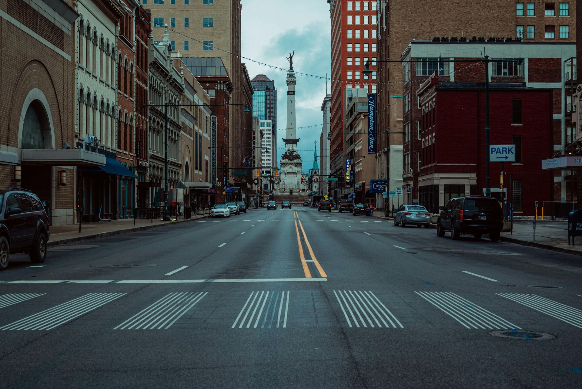 Empty city street lined with brick buildings, leading to a distant tower under a cloudy sky.