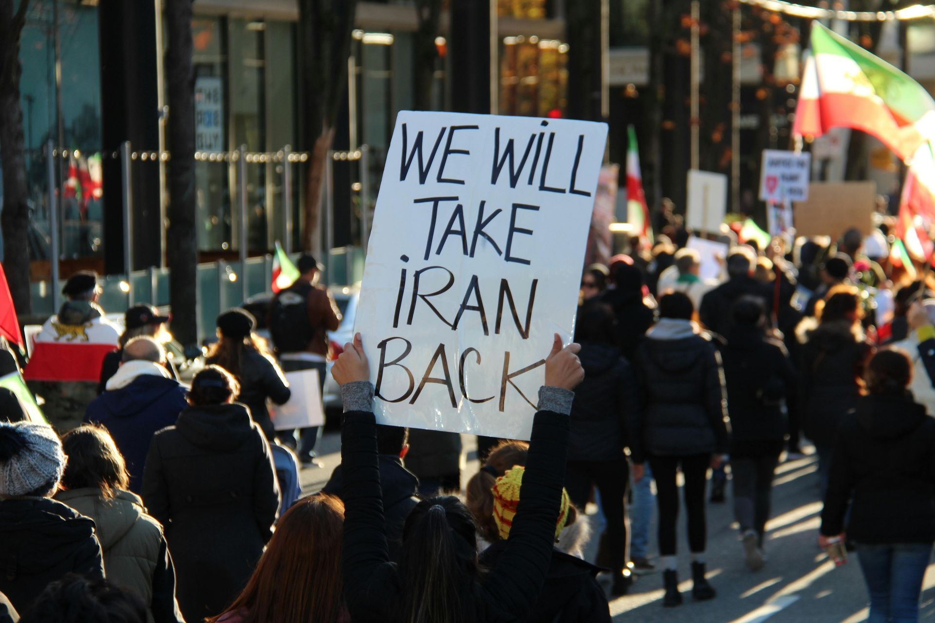 Crowd marching on a city street with a sign reading “WE WILL TAKE IRAN BACK.”