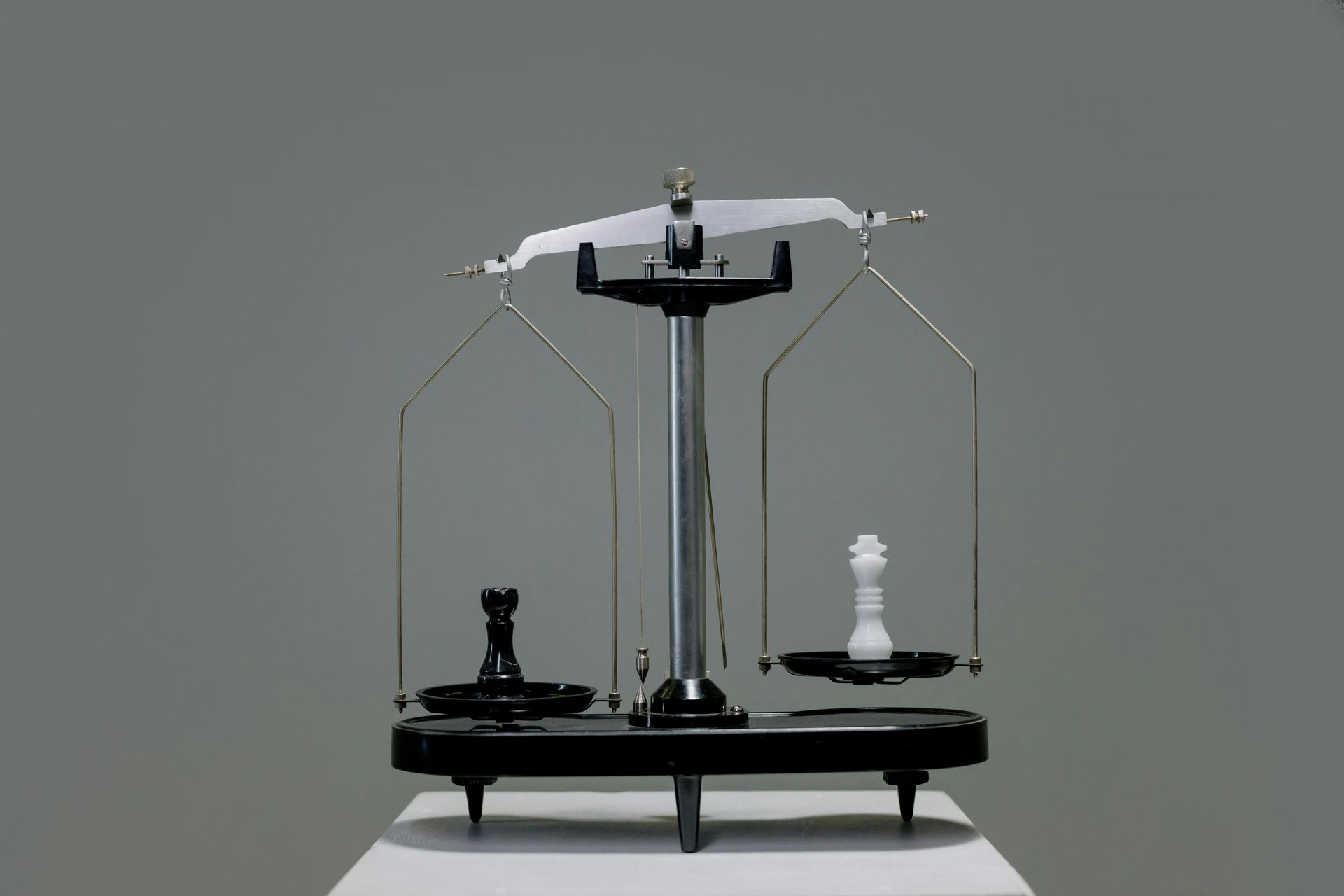 A balance scale with a black rook and a white chess king on each side, set against a gray backdrop.