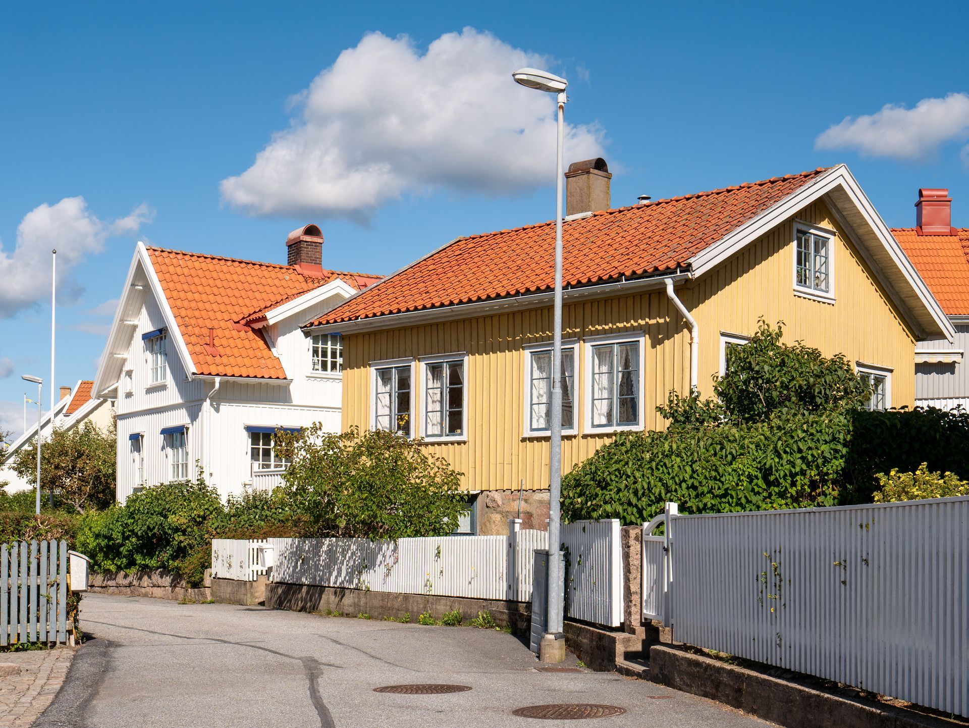 Yellow and white houses with orange roofs line a street under a blue sky, white picket fence.