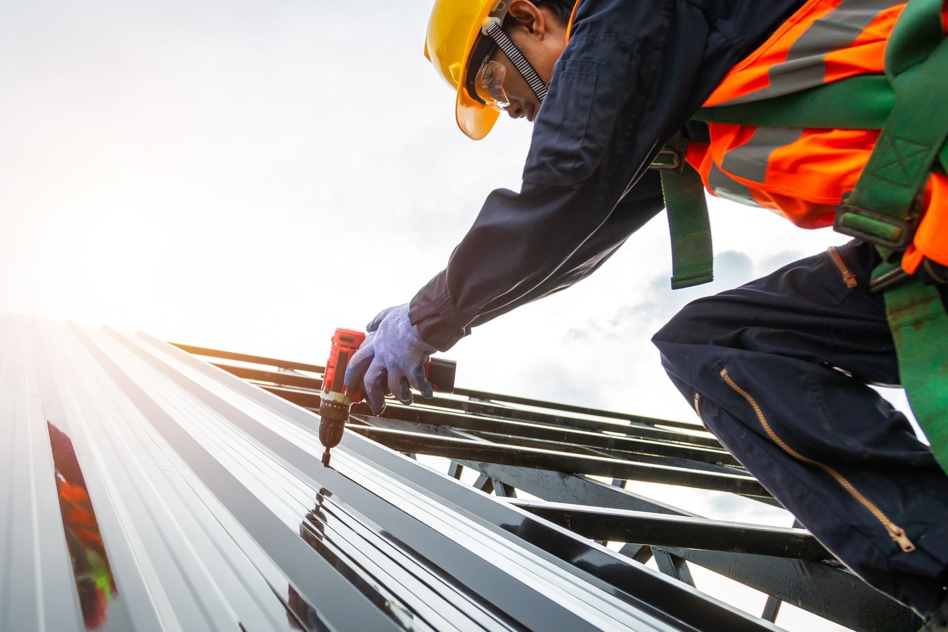 Roofer in orange vest and hard hat using a drill on a metal roof. Bright sunlight.