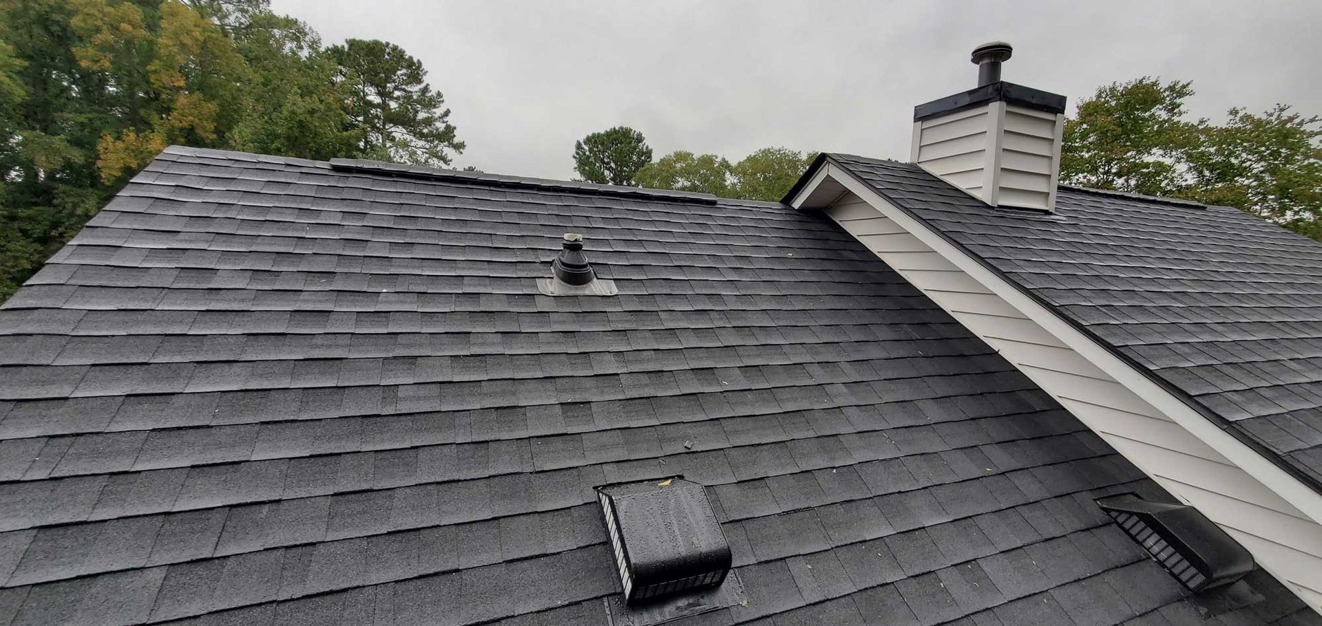 A dark gray asphalt shingle roof with a white chimney against an overcast sky, trees in the background.