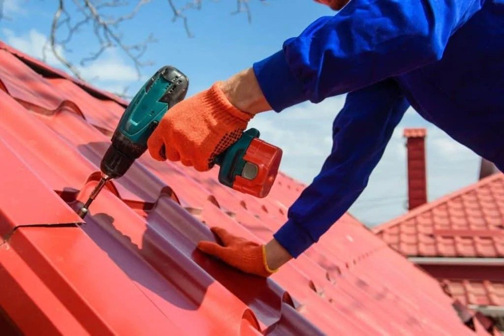 Person in blue work clothes, orange gloves, using a drill to install red metal roofing on a house. Person in blue work clothes, orange gloves, using a drill to install red metal roofing on a house.
