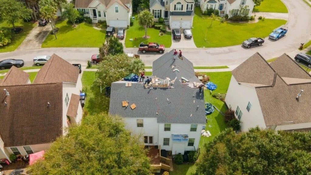 Aerial view of workers replacing a gray shingle roof on a two-story white house in a neighborhood.