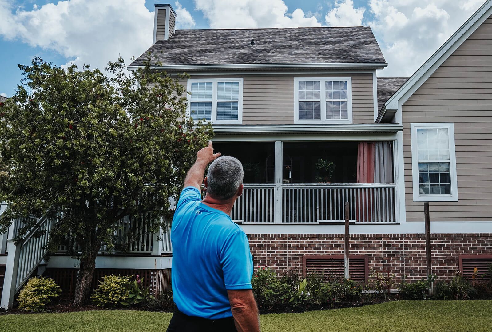 Man in blue points up at two-story house with porch and gray roof on a sunny day.