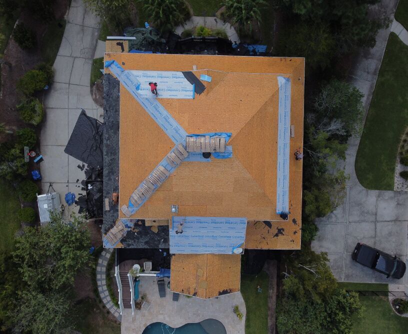 Overhead view of a house roof under construction. Workers are applying new shingles; debris and tools are scattered around.