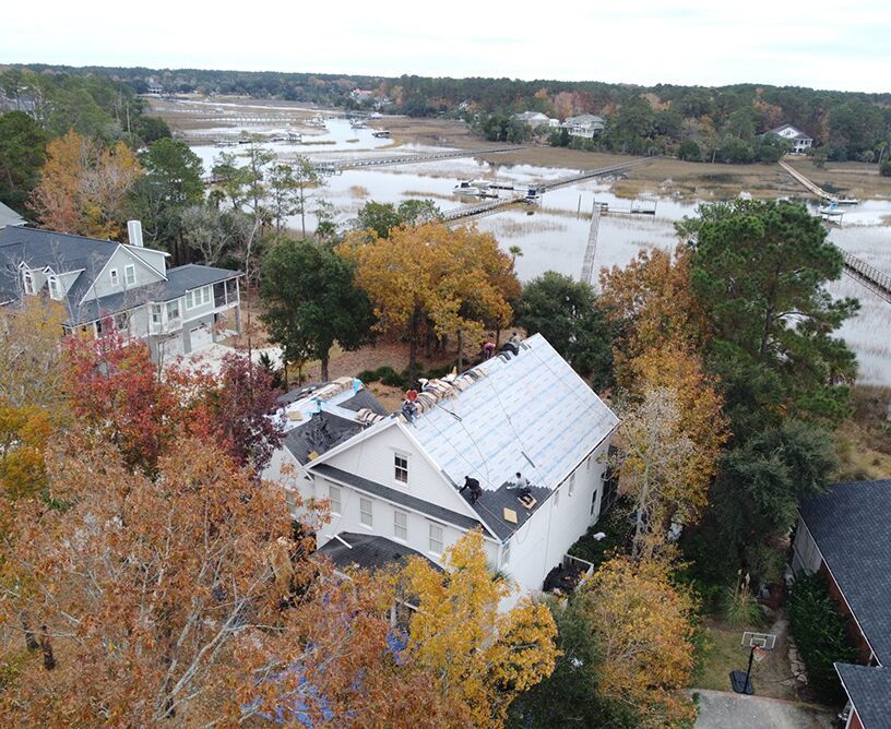Aerial view of a white house with roof repairs, surrounded by colorful trees, near a body of water.