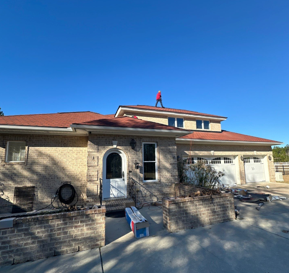 Person on a roof of a brick house with red roof shingles, sunny day.