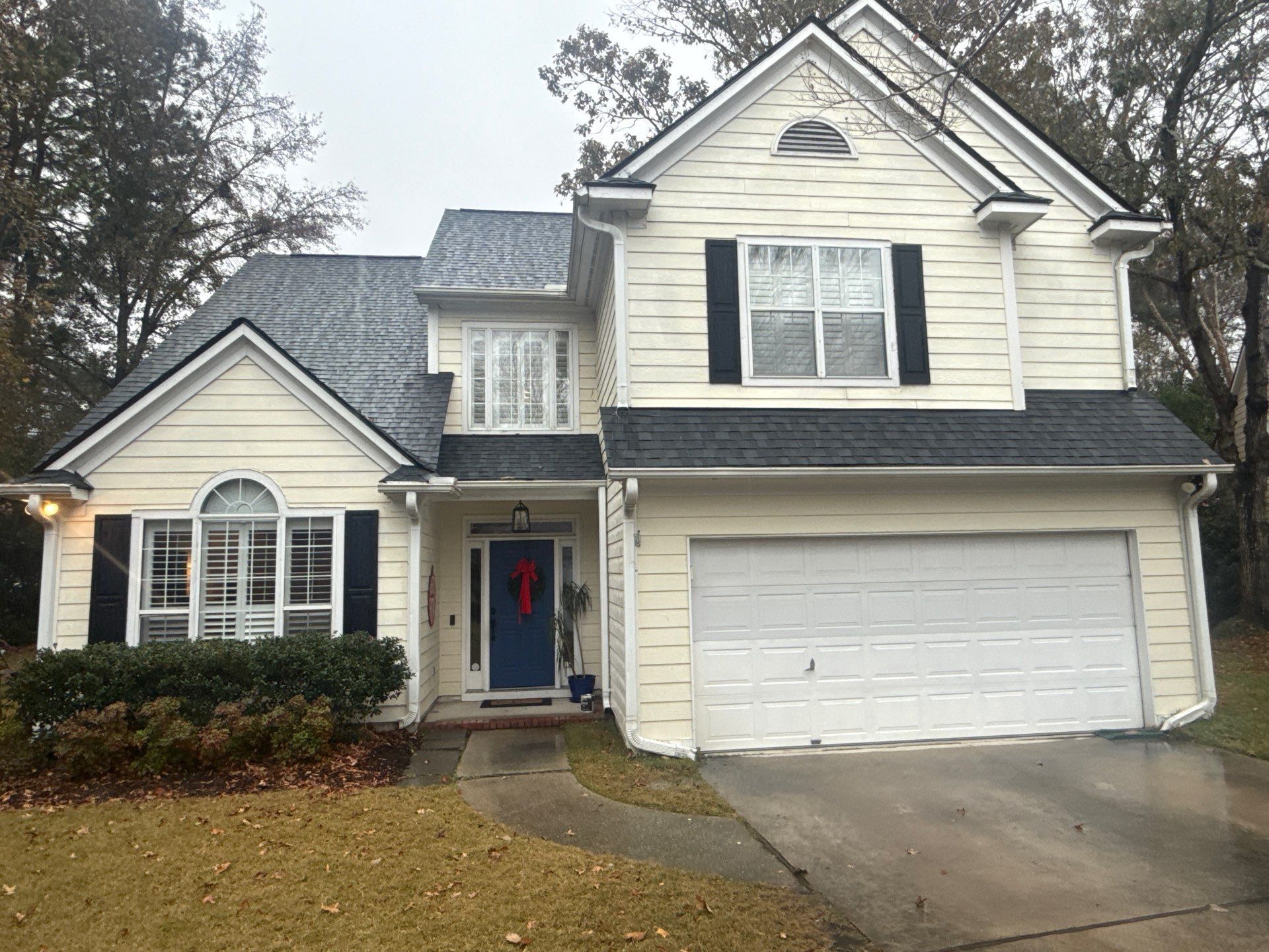 Two-story house with tan siding, black shutters, and a dark gray roof. A blue front door has a wreath.