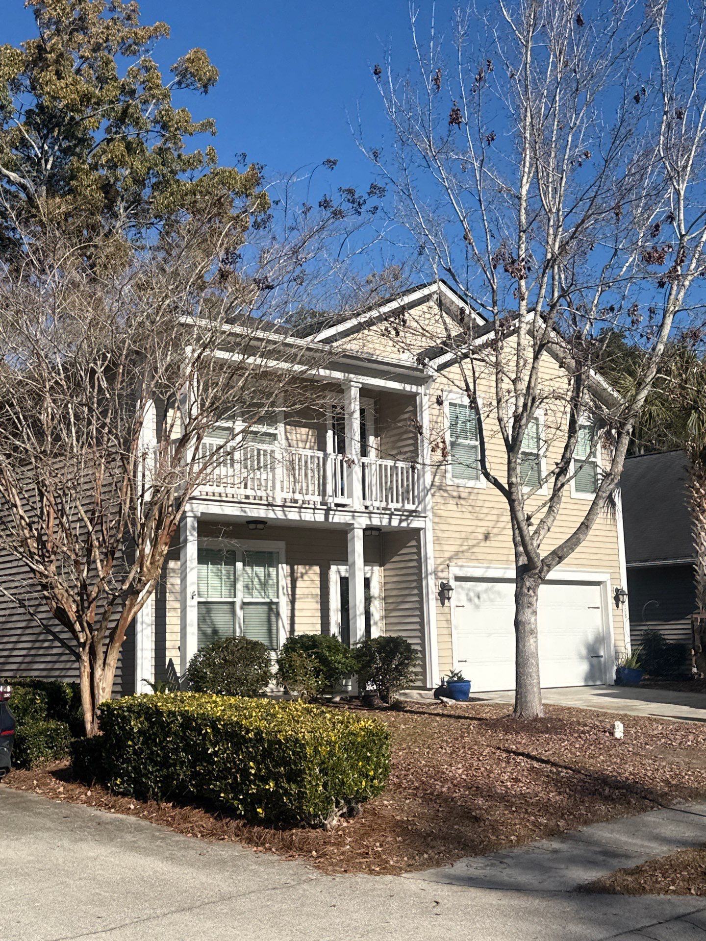 Two-story tan house with white balcony, shrubs, bare trees, and blue sky.