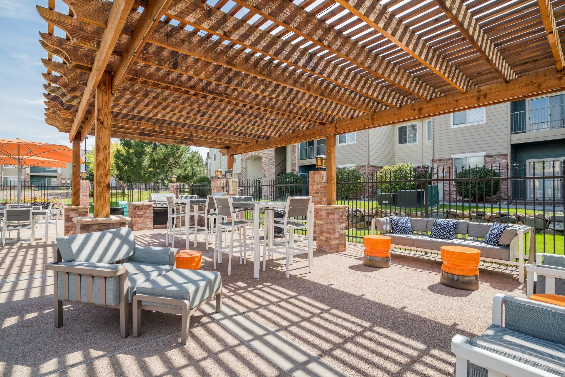 Outdoor patio with seating under a wooden pergola, includes bar, sofas, and tables, with buildings in the background.