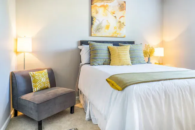Bedroom with a bed, chair, two lamps, and yellow and gray accents.