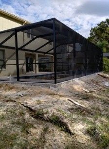 Black mesh screened patio enclosure beside a house on a dirt lot under a cloudy sky