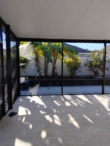 Covered patio with black mesh screens, sunlit concrete floor, and a view of palm trees and hills beyond the fence