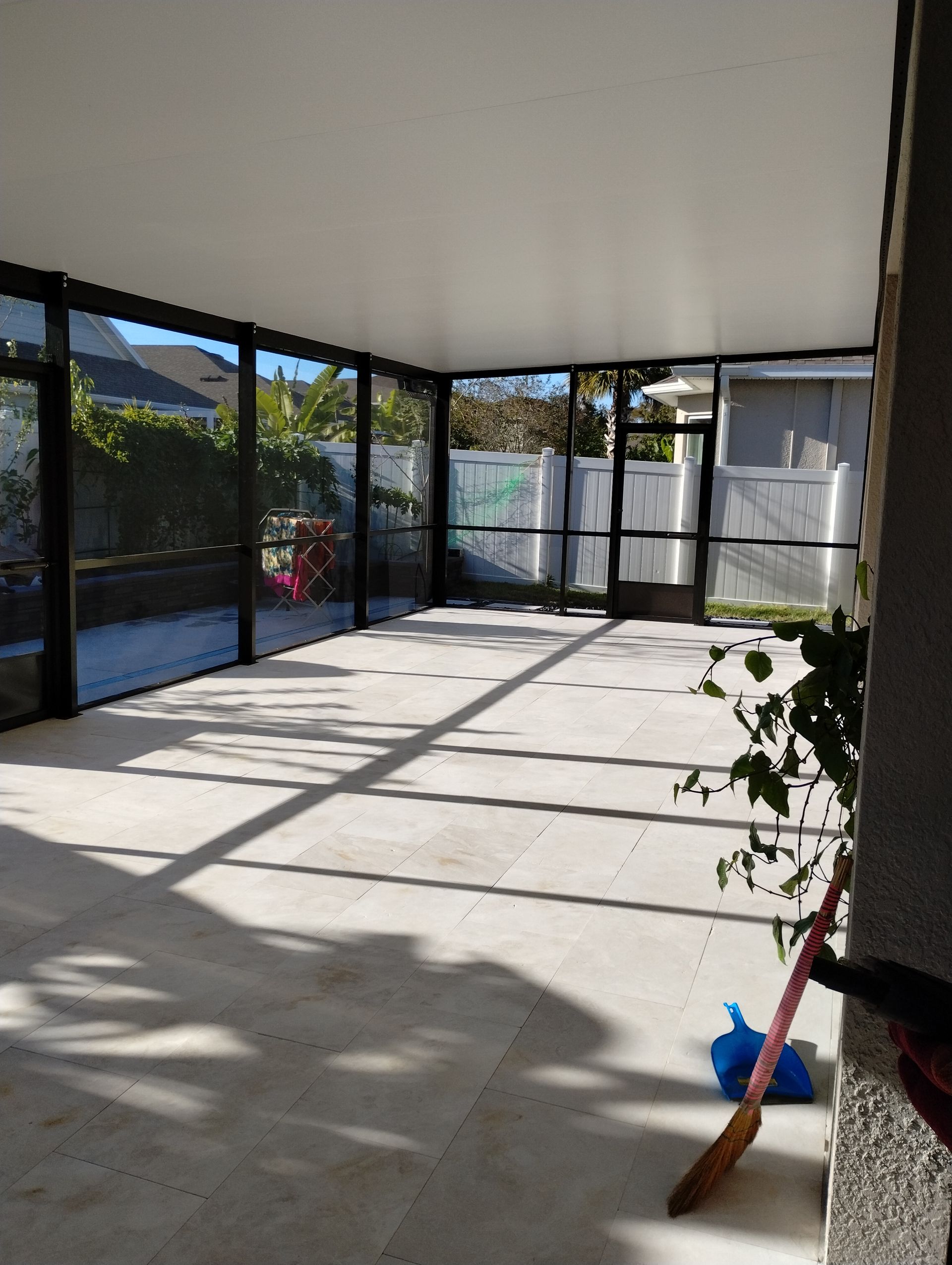 Empty sunroom with tile floor, large windows, and sunlight casting shadows; broom and dustpan in corner