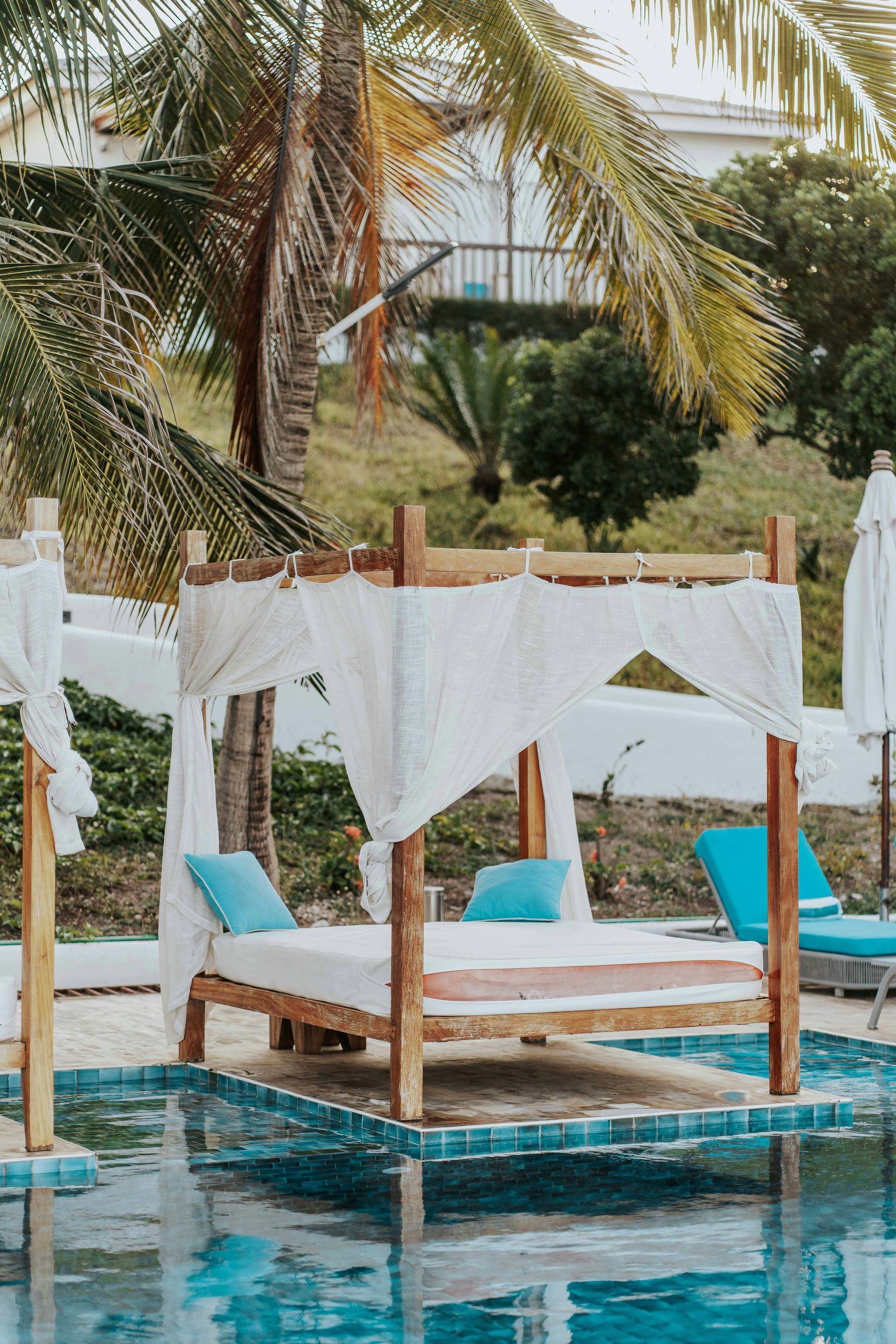 Poolside daybed with white drapes, teal cushions, and palm trees in a tropical resort setting