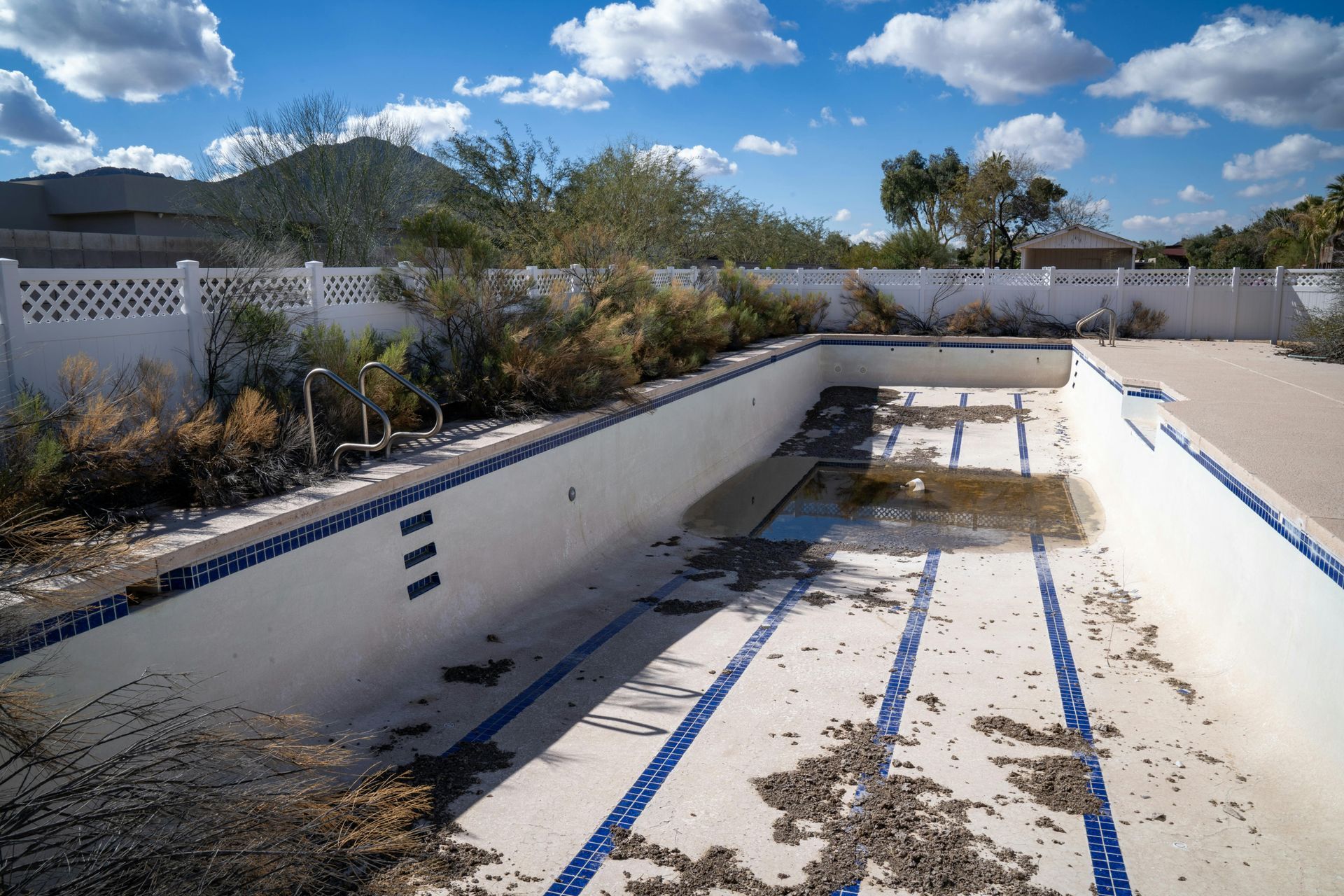 Empty outdoor swimming pool with peeling white walls, blue lane lines, and overgrown debris under a cloudy sky