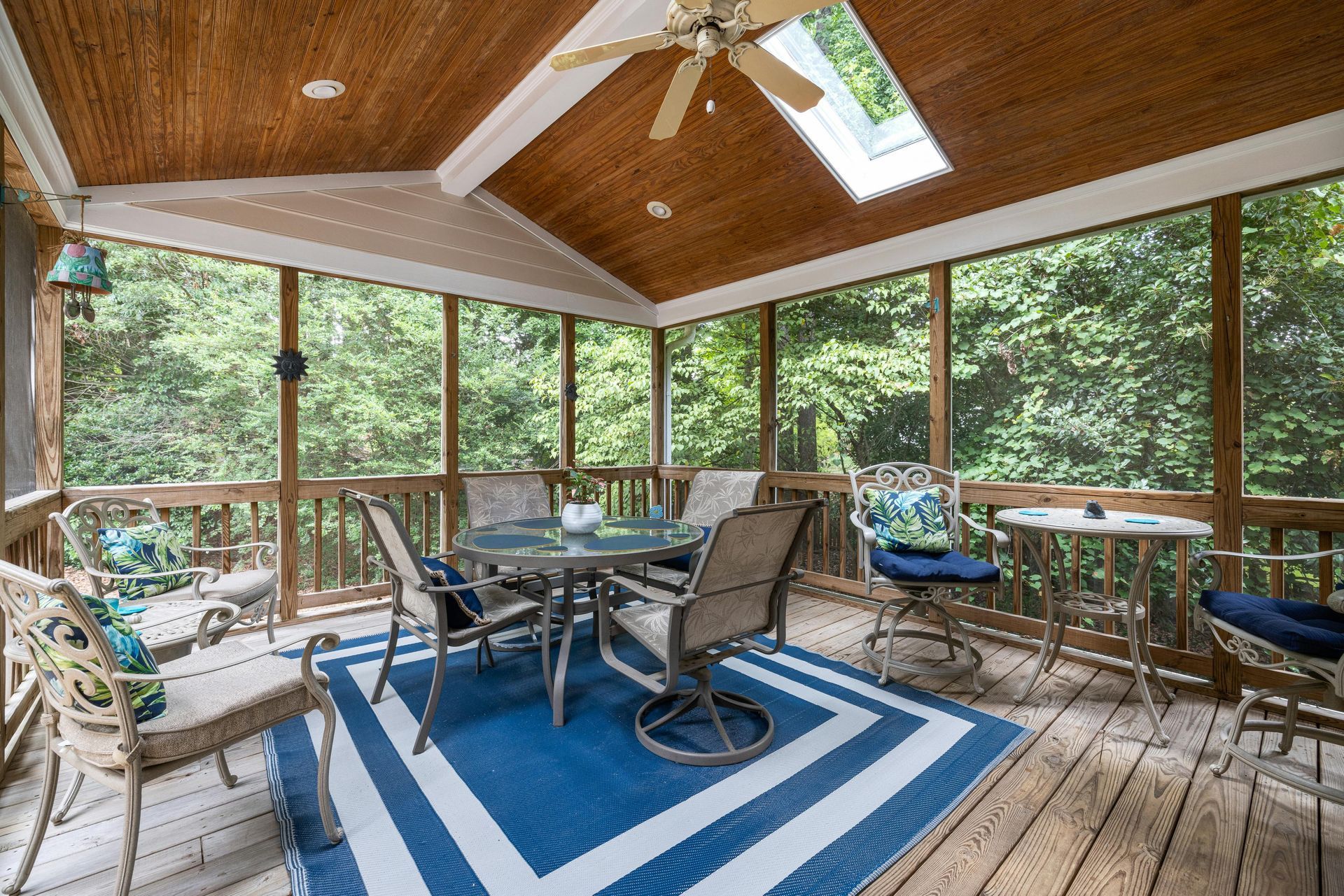 Screened porch with wood ceiling, blue rug, patio furniture, and wooded view through windows