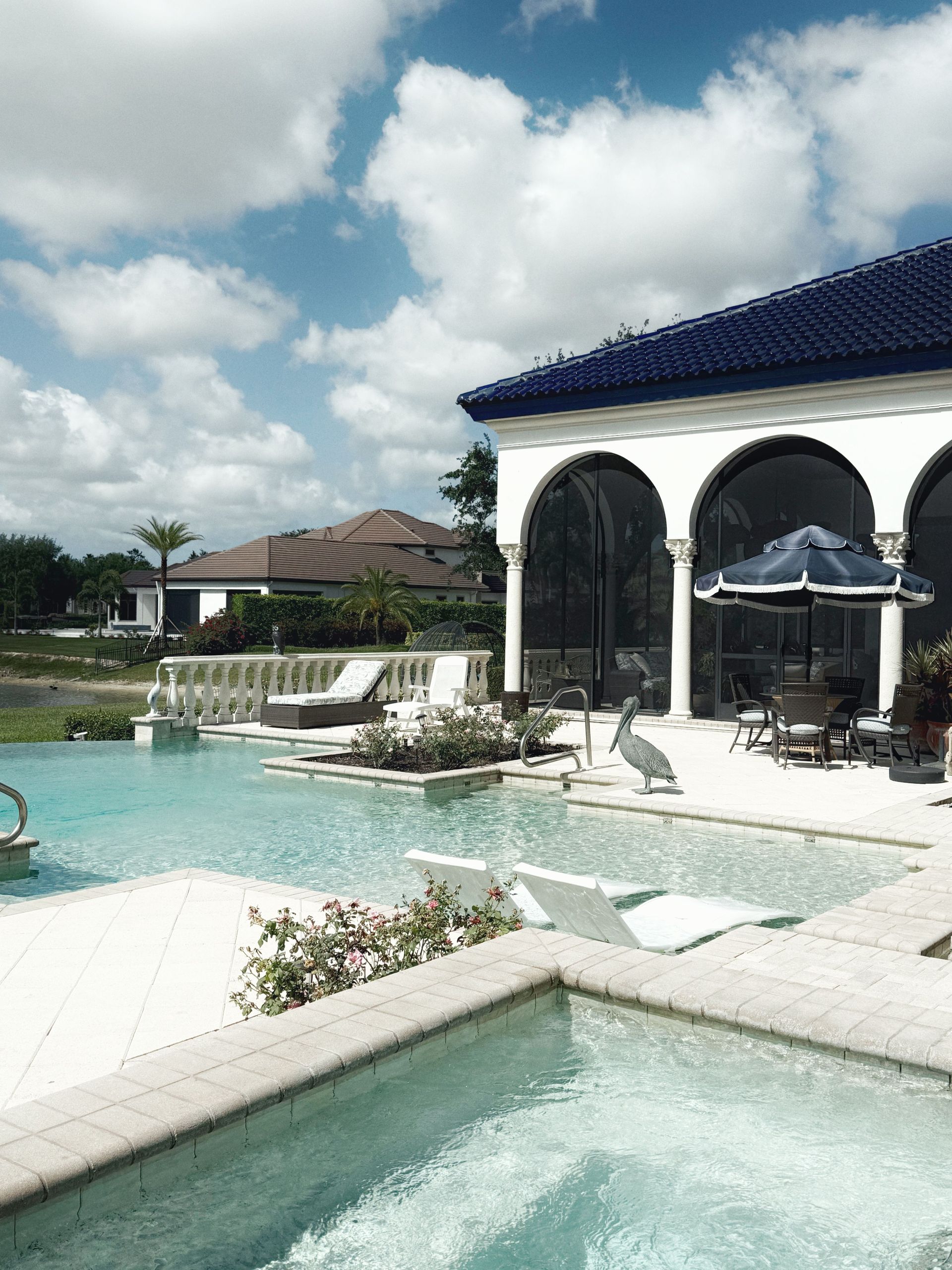 Luxury poolside resort with white lounge chairs, arched patio, and blue-tiled roofs under a cloudy sky