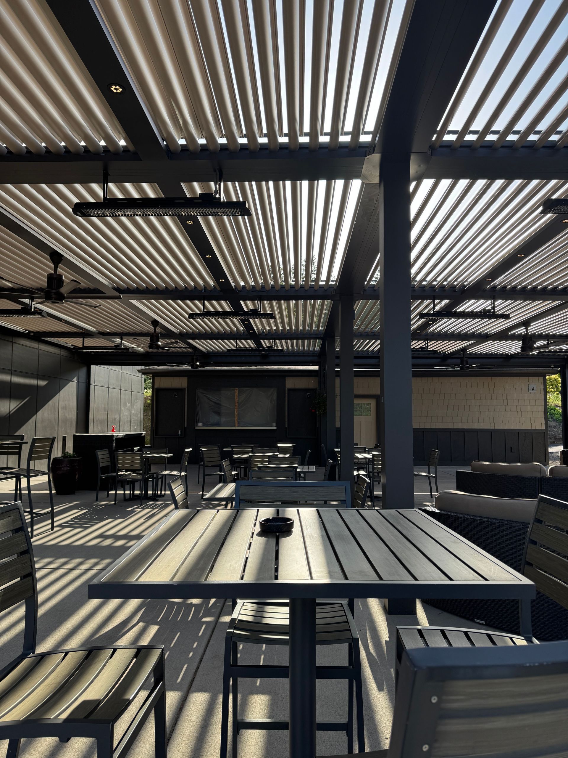 Outdoor patio seating under a slatted roof with tables, chairs, and a large pillar in shade.