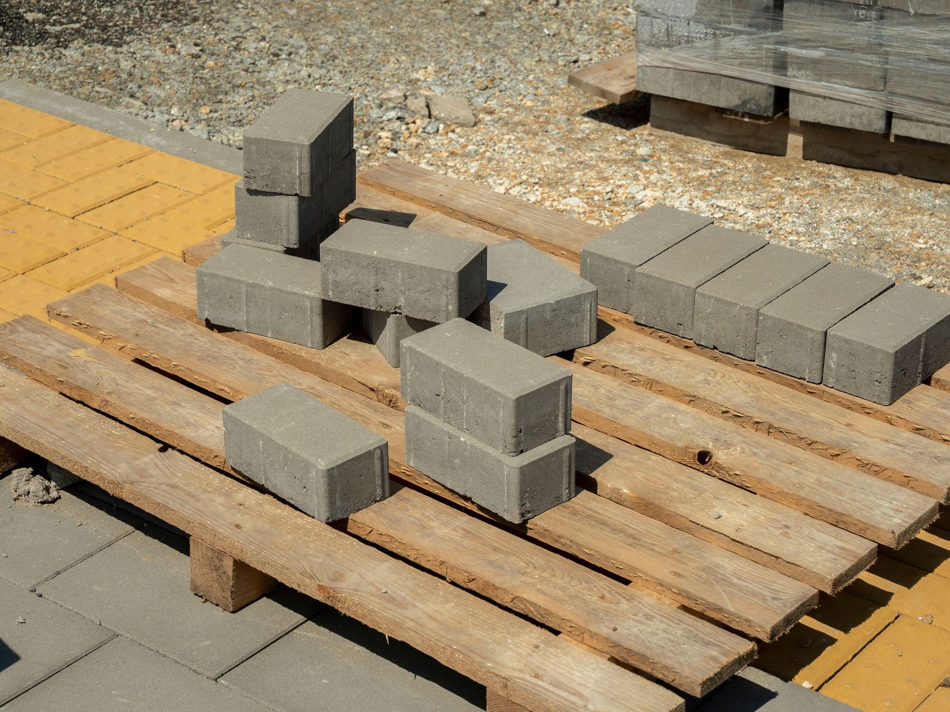 Stacks of gray paving blocks on wooden pallets at a construction site, with yellow tiles nearby.