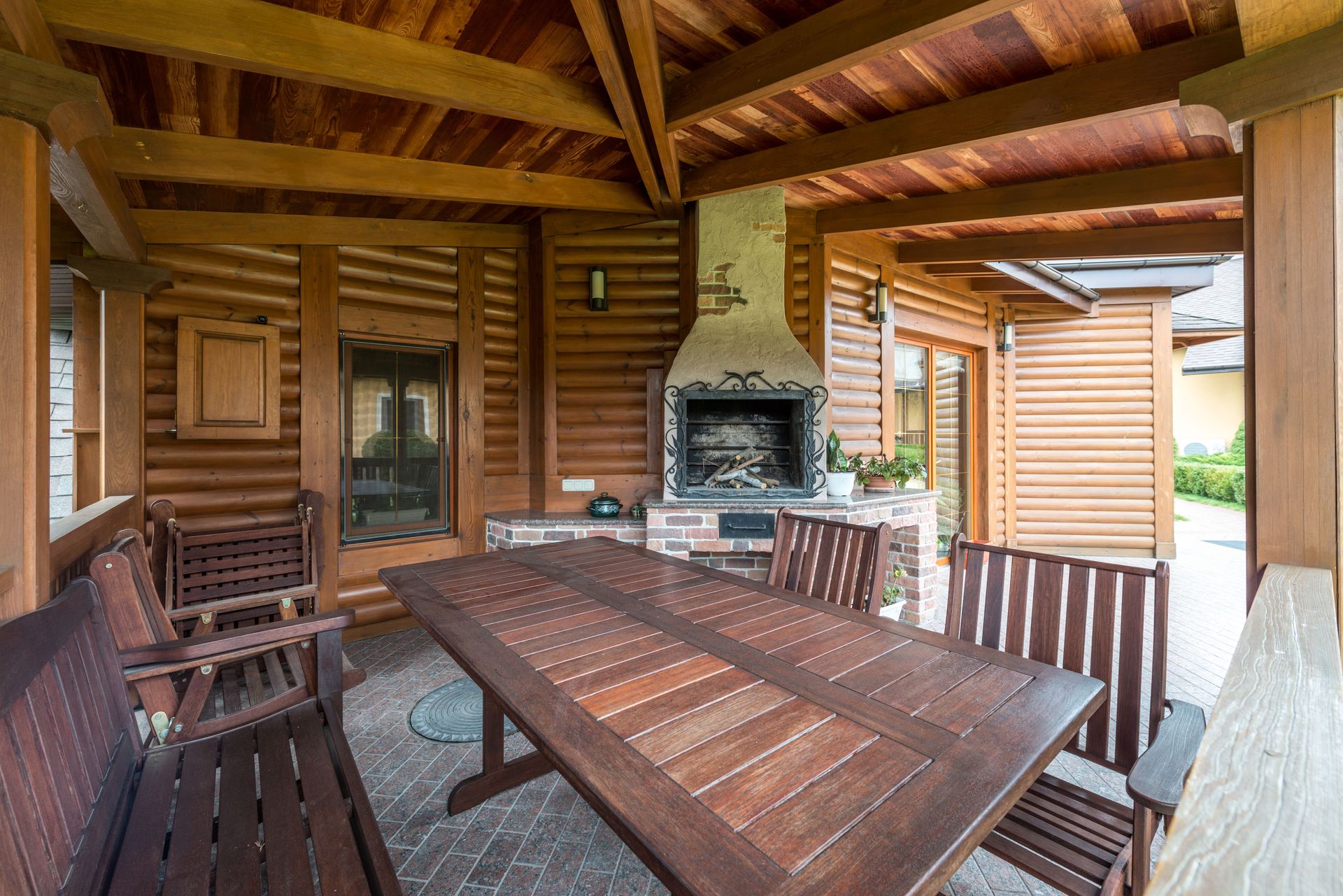 Covered wooden patio with picnic table, benches, and stone fireplace in a rustic cabin setting