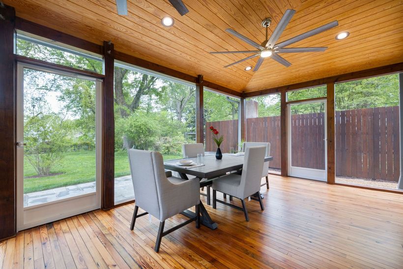 Sunlit dining nook with glass walls, wooden ceiling, gray chairs, and forest view