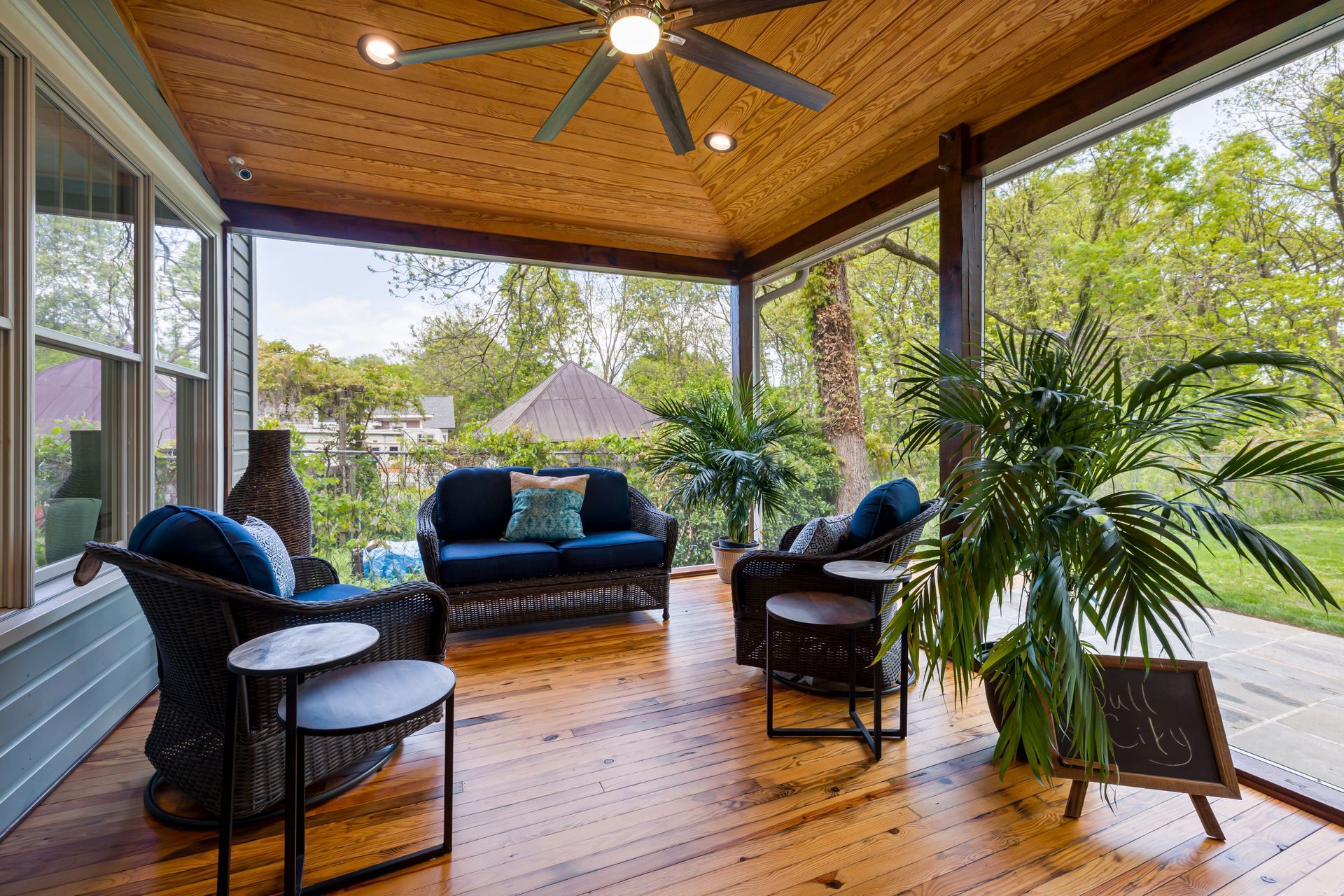 Covered patio with wicker seating, blue cushions, wood ceiling, and lush garden view