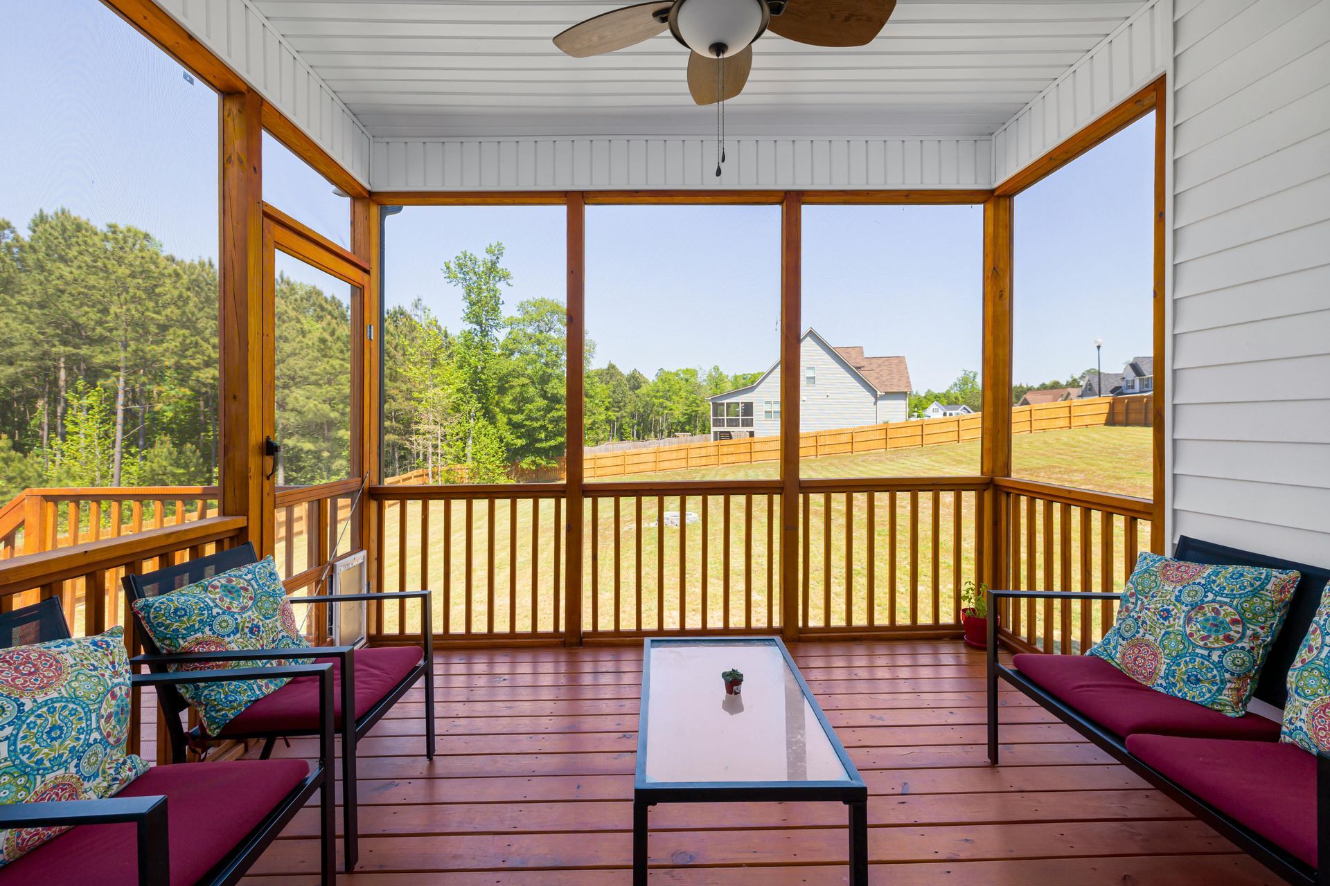 Covered porch with red floor, wicker chairs, coffee table, and a view of green fields and houses.