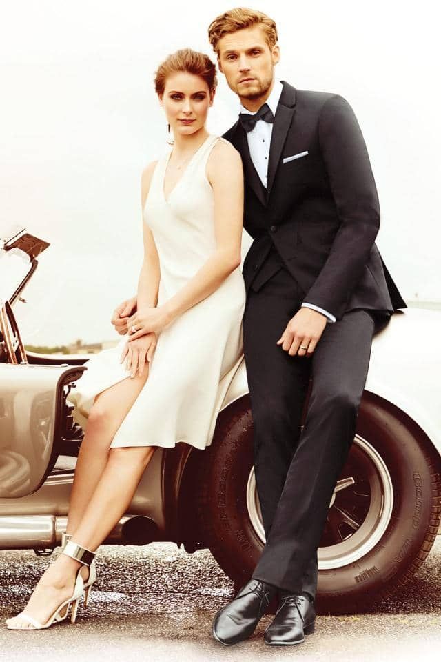 a man in a tuxedo and a woman in a white dress are standing next to a car .