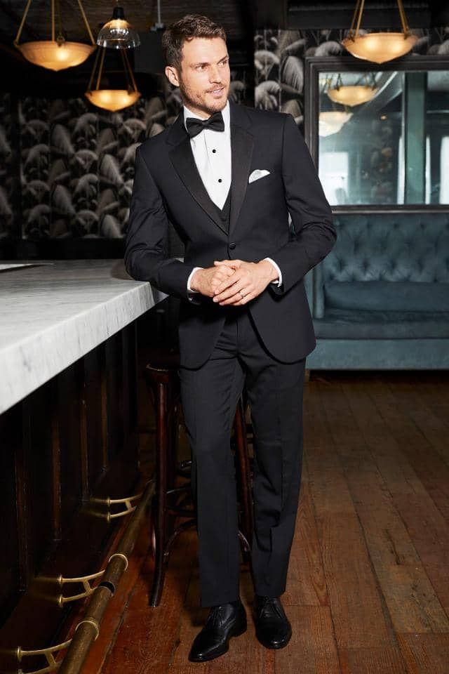 a man in a tuxedo and bow tie is standing next to a bar .