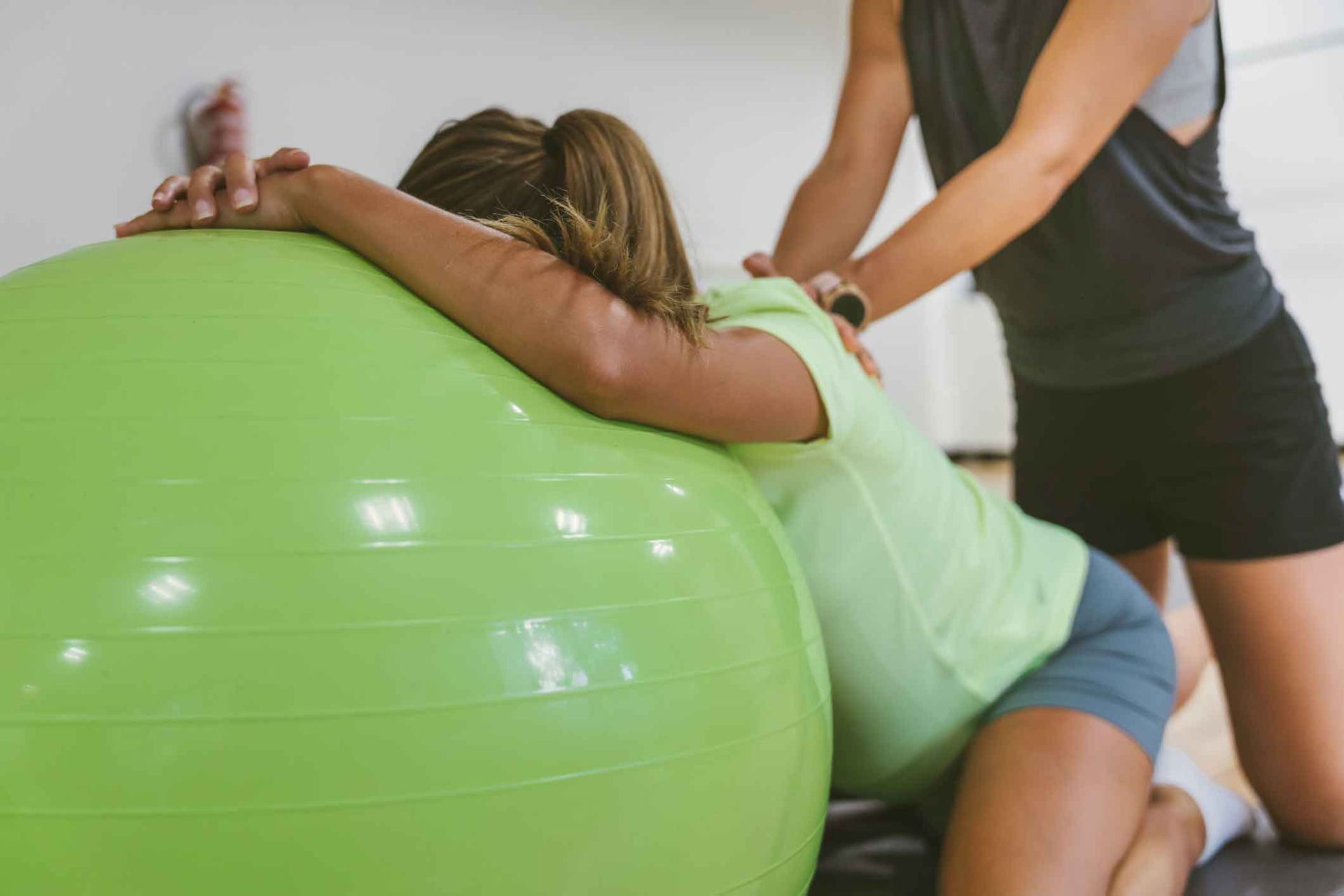 A woman is laying on a green exercise ball while a man helps her.