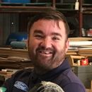 A man with a beard is smiling and holding a tool in a warehouse.