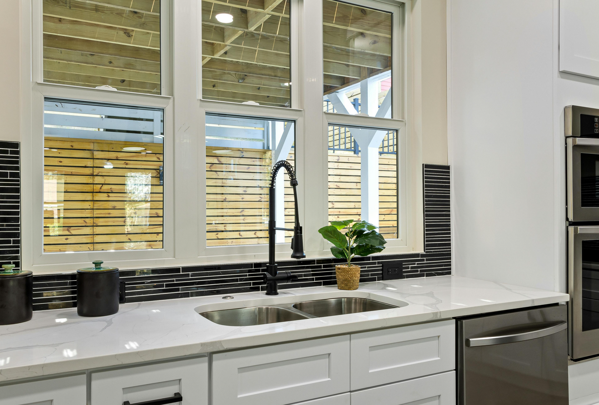 Modern kitchen with white cabinets, marble countertop, black faucet, and window overlooking a wooden deck.