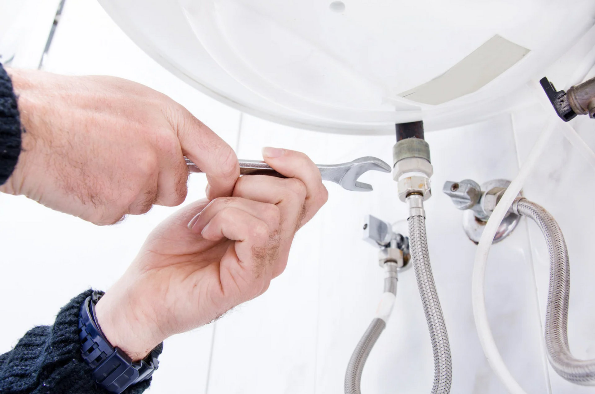 Person using a wrench to work on plumbing pipes connected to a water heater in a white-tiled space.