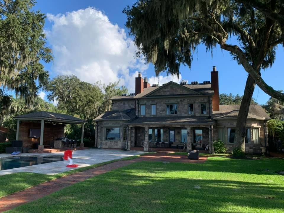 Stone mansion with pool, patio, and lawn, under a sunny sky with Spanish moss-covered trees.