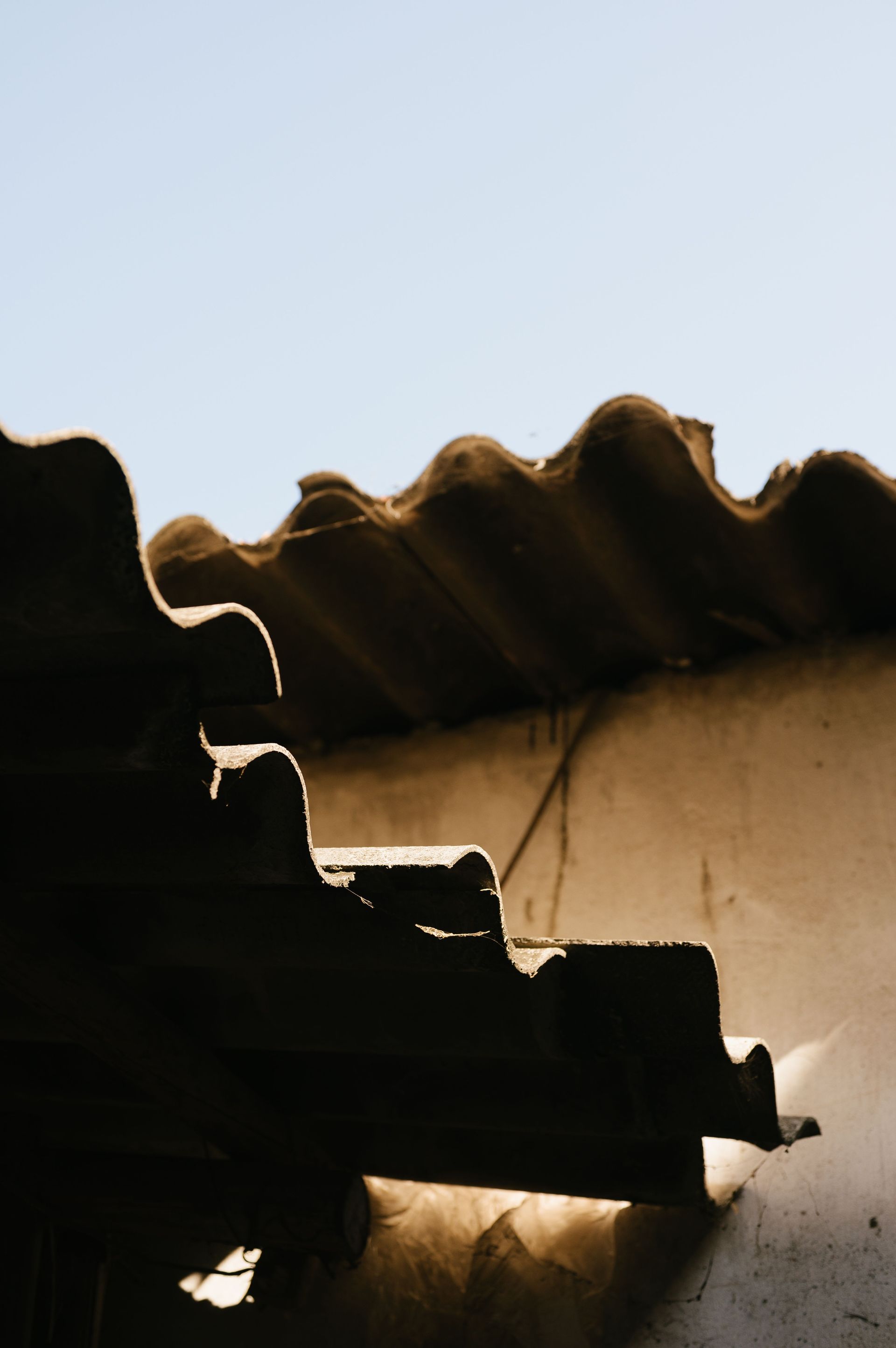 Sunlit corrugated roof edge against a pale blue sky, with a beige wall below.
