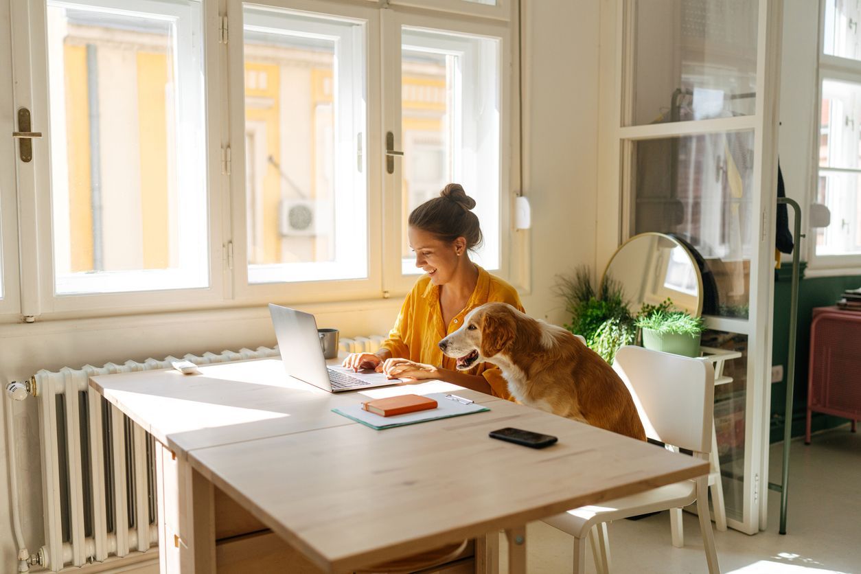 A woman is sitting at a table with a dog and using a laptop.
