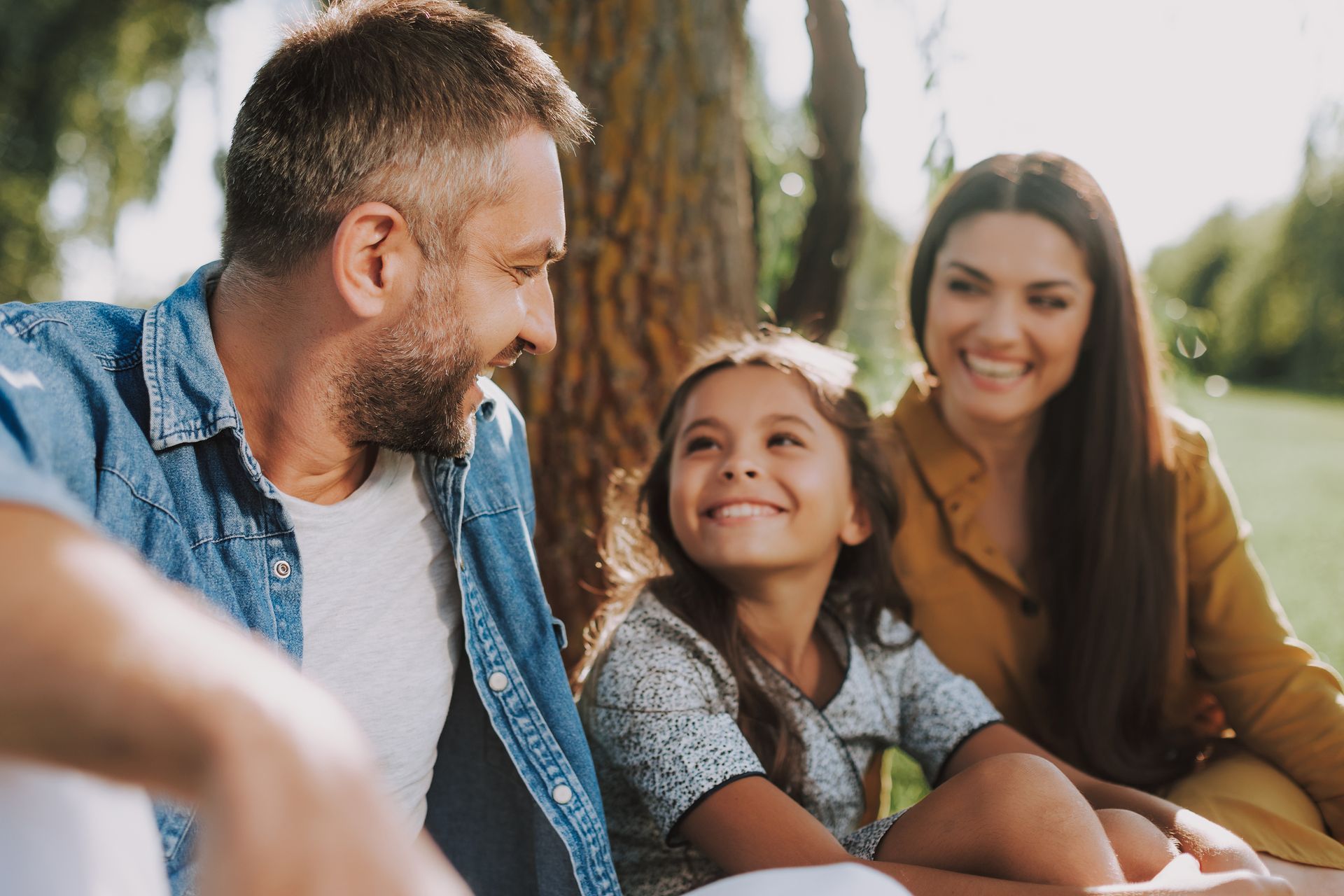 Family of three smiles together under a tree in a sunny park.