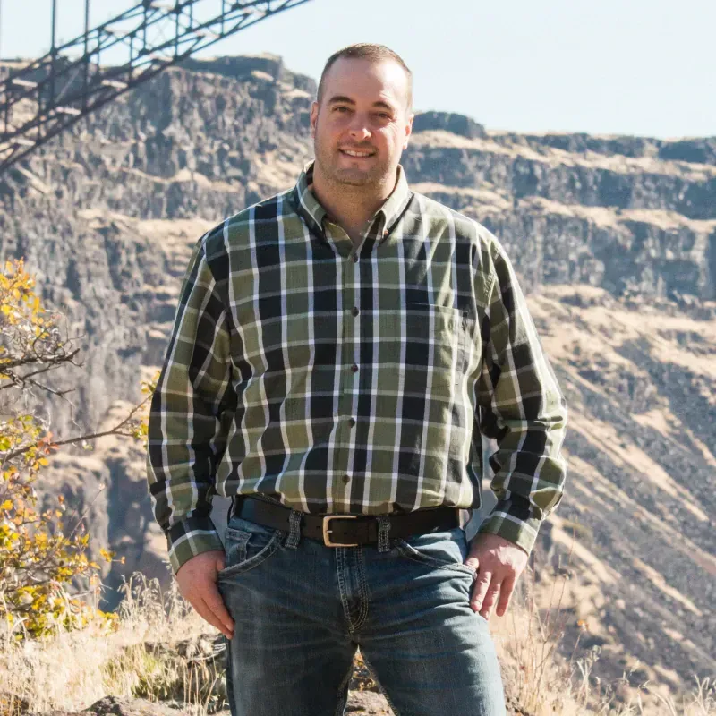 A man in a plaid shirt and jeans is standing in front of a mountain.