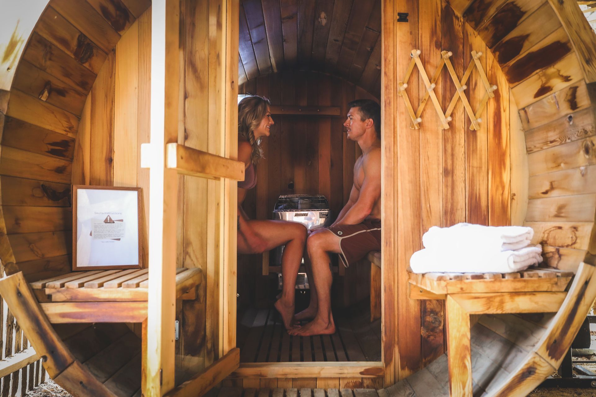 Two people sit inside a small wooden sauna, facing each other, with towels nearby.