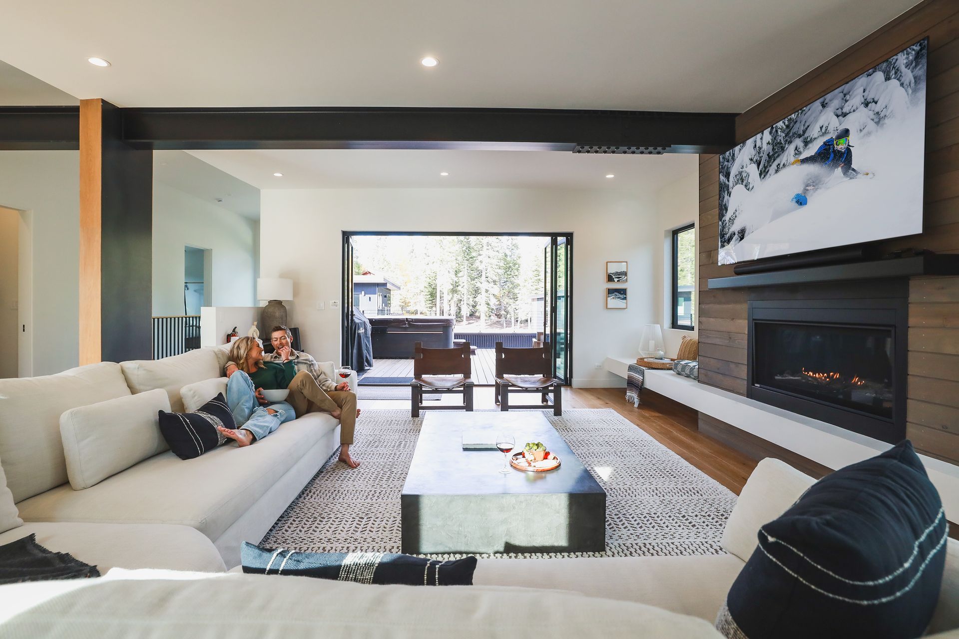 Modern living room with people on a sofa, fireplace and TV, open to a bright outdoor patio.