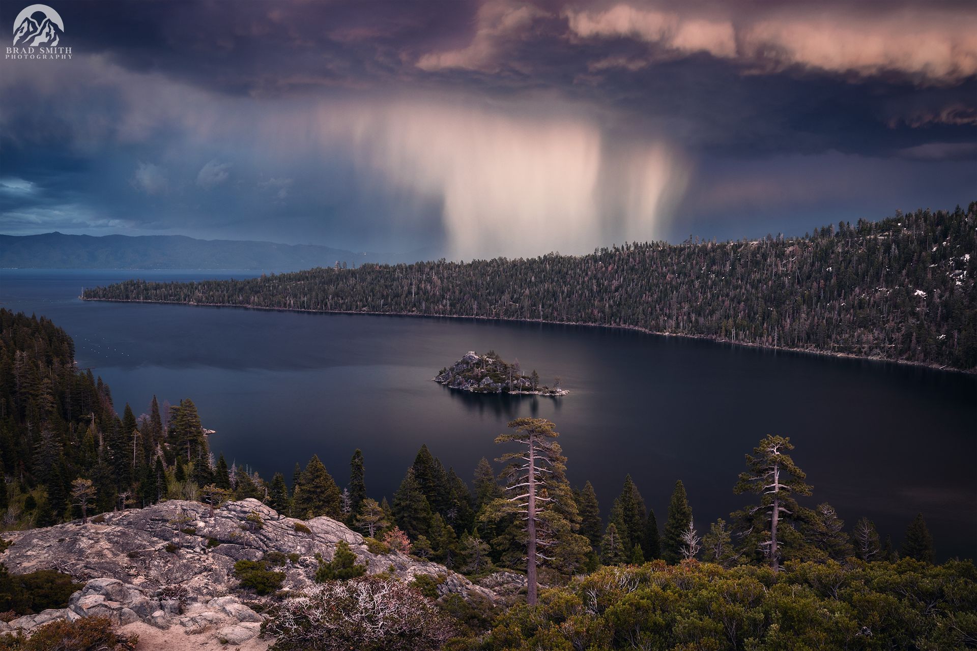 Stormy lake with a small island, dark forested shoreline, and a pink sunset sky