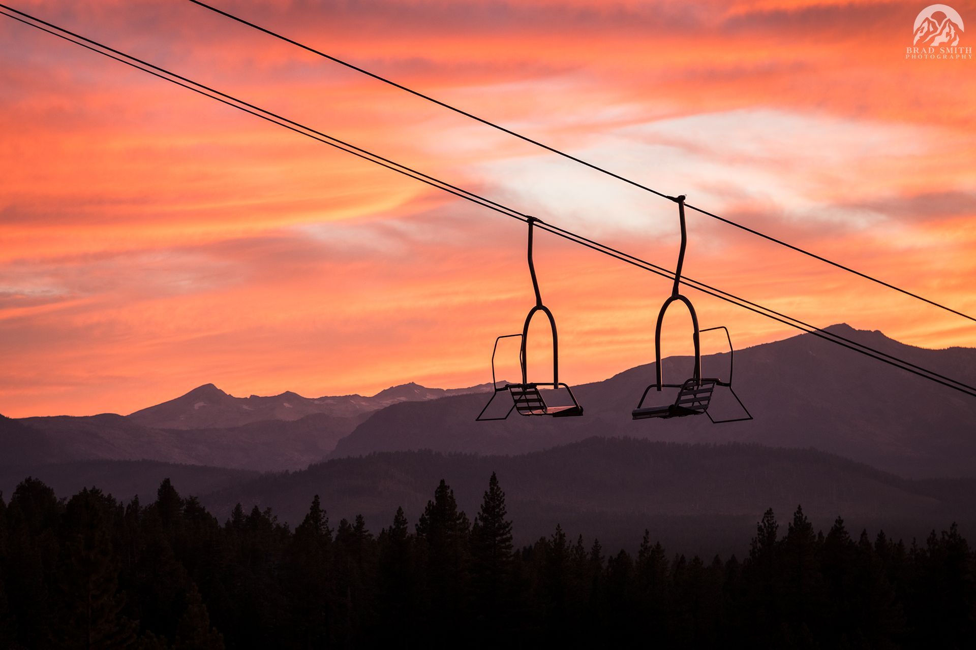 Ski lift chairs silhouetted against a pink-orange sunset over mountains and pine trees