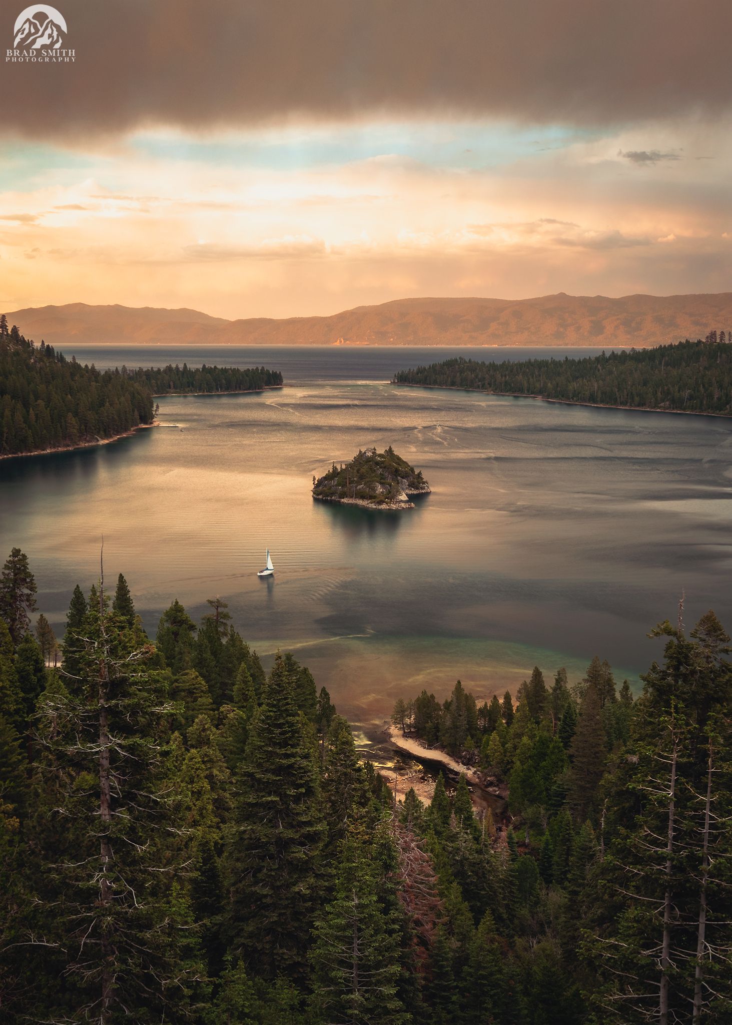 Sunset over a calm lake with a small island, pine forest shoreline, and distant hills