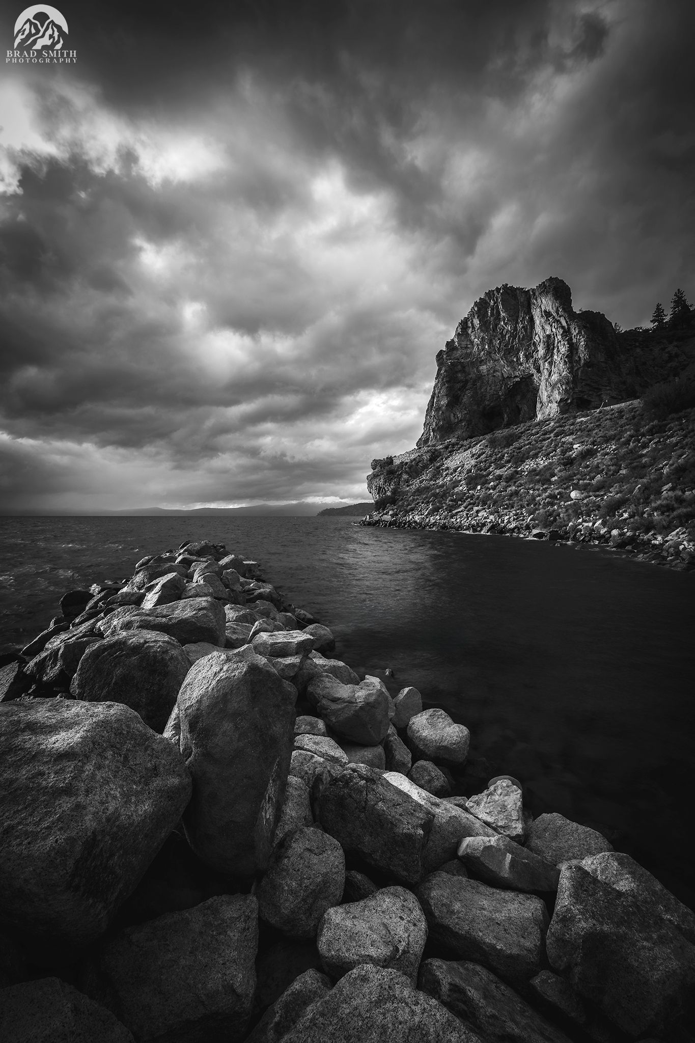 Rocky coastline under dramatic cloudy sky, with dark water and cliffs in black and white.