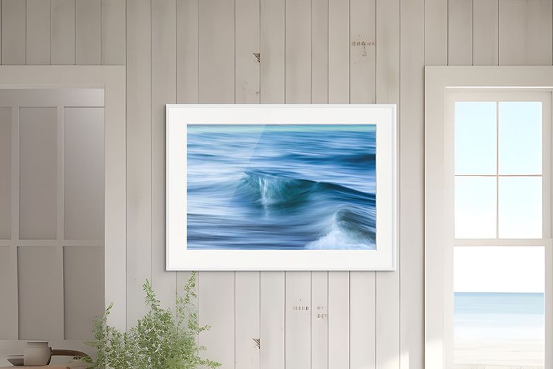 Framed blue ocean wave painting on a light wood wall beside a bright window