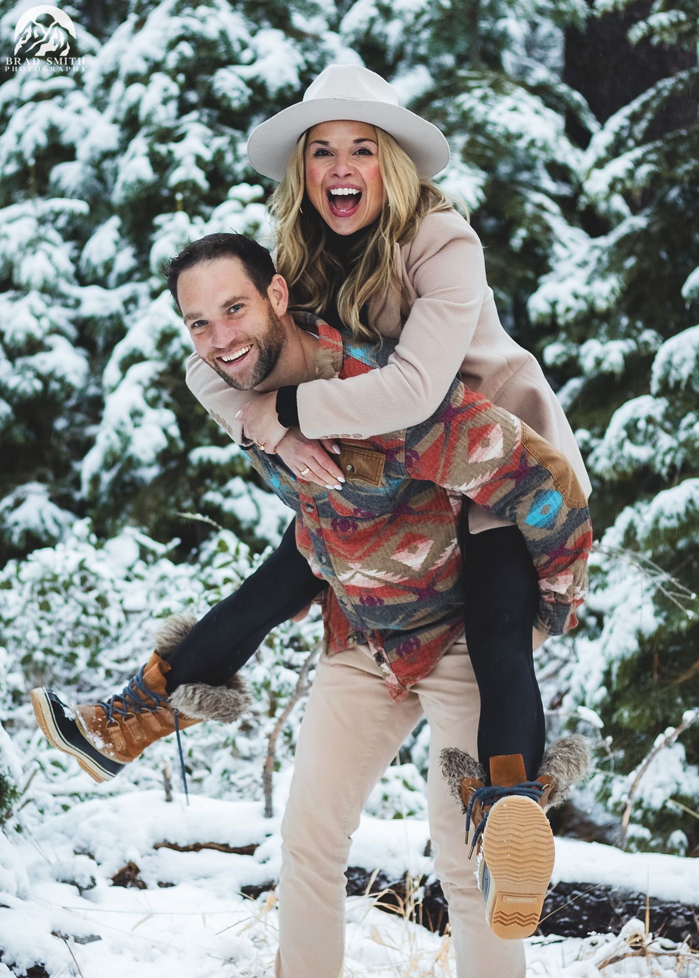 Man giving a woman a piggyback ride in a snowy forest, both smiling and wearing winter clothes